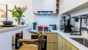 Een compacte keuken met witte kastjes, een zwarte koffiezetapparaat, een rode pan en een kleine eethoek met twee stoelen.