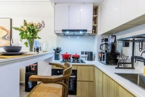 A compact kitchen with white cabinets, a black coffee machine, a red pot, and a small dining area with two chairs. - Sky Love Lavington – Bright & Stylish 1BR Retreat with Wi-Fi & Smart TV (Nairobi)