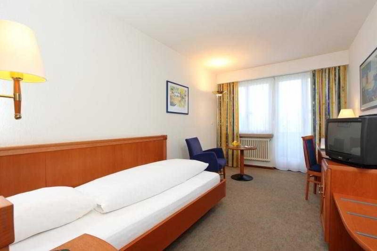 A hotel room with a bed, a desk with a television, a chair, and a window with curtains.