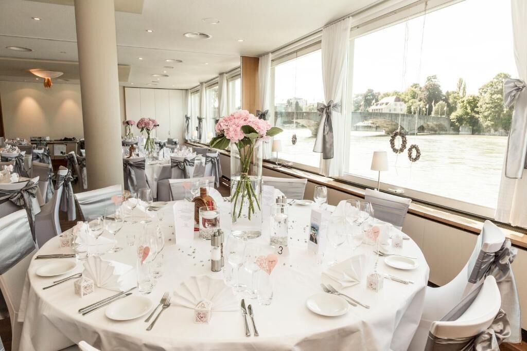 Una sala de banquetes amb taules rodones preparades per a un esdeveniment formal, grans finestres amb vista al riu i un penjador de cristall al sostre.