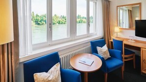Ein Hotelzimmer mit großem Fenster, einem runden Tisch, blauen Stühlen, einem Holzbüro, einem Fernseher und einem Spiegel.