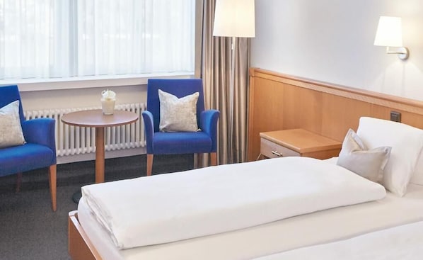 A hotel room with a bed, two blue armchairs, a small table with a candle, a lamp, and a view of the outdoors through a window. - Hotel Schiff am Rhein (Rheinfelden)