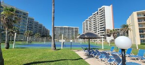 A tennis court surrounded by palm trees and lounge chairs, with high-rise buildings in the background. - Sleeps 10 Direct Beach Heated Pool (South Padre Island)