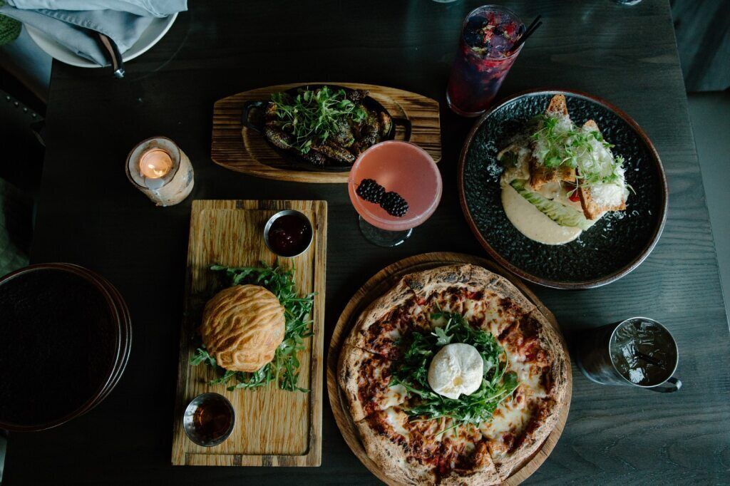 Een eettafel met diverse gerechten, waaronder pizza, hamburger en salade, samen met drankjes en een kaars.