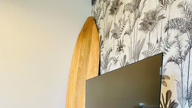 Uma sala de estar moderna com uma TV de tela plana sobre um suporte de madeira, um banquinho trançado e um vaso verde no chão.