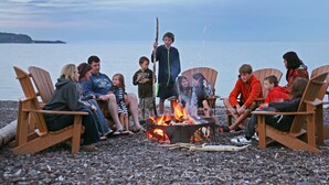 A group of people sitting on wooden chairs around a campfire on a pebbled beach with a body of water and a distant landmass in the background. - Lakeview At The Lodge Two Bedroom Suite (Two Harbors)