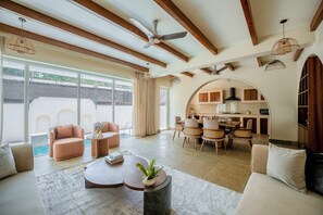 A spacious living area with a large window, a ceiling fan, and a kitchen visible through an archway. - Staymaster White Castle-3BHK | Pvt Pool | Vagator  (Vagator)
