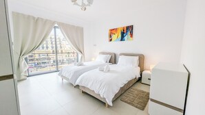 Una camera d'albergo con due letti, un'ampia finestra con tende, un quadro appeso al muro e un comodino con una lampada.