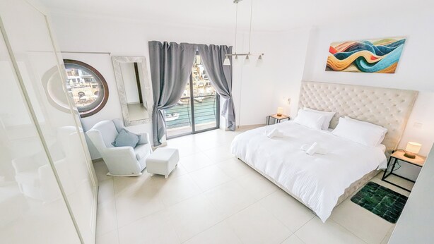 Una moderna camera d'hotel con un letto grande, una zona salotto con una poltrona e un tavolino, una finestra ampia con tende, e un quadro astratto dai colori vivaci appeso al muro.
