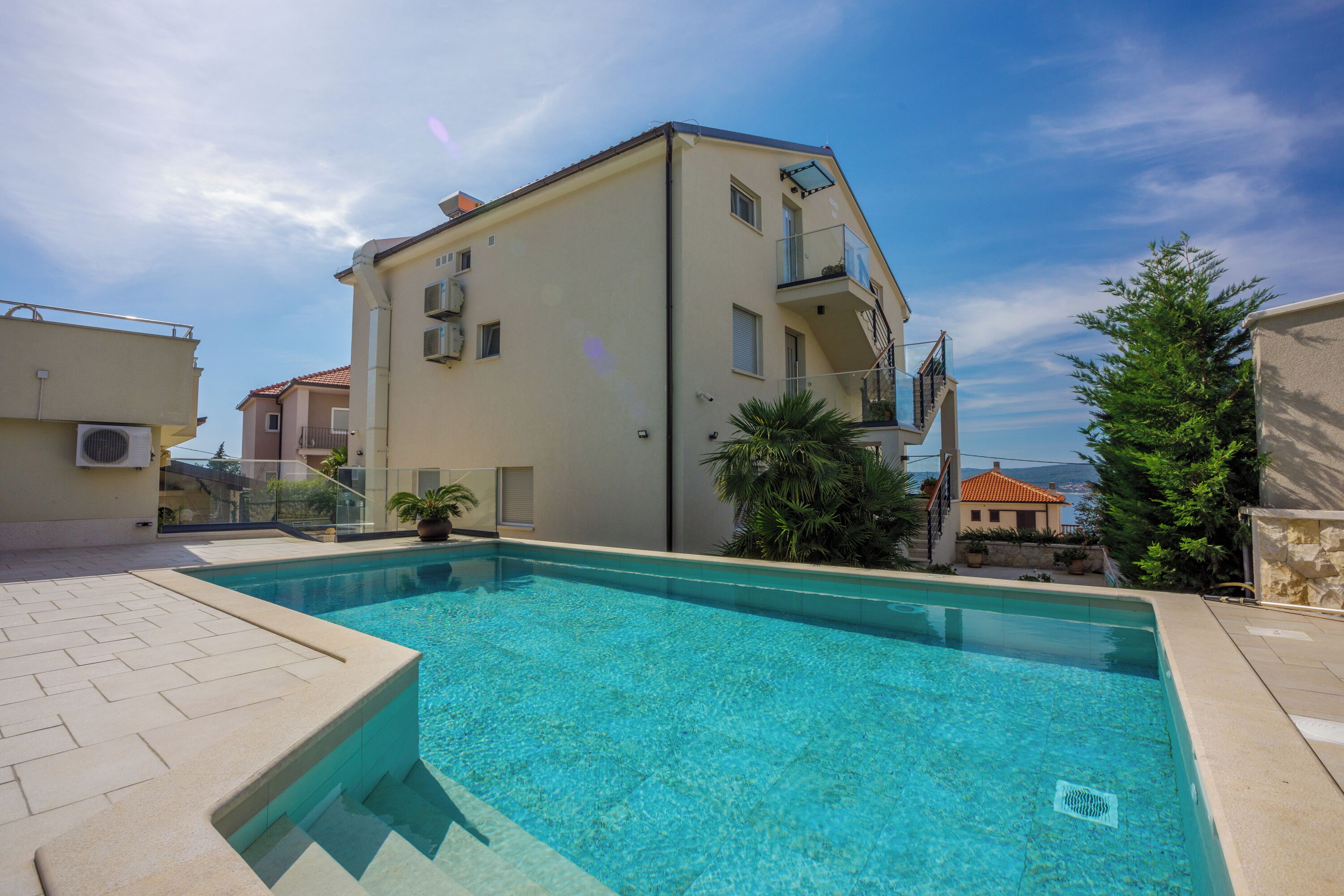 A swimming pool with a clear blue water, surrounded by a building with a balcony and a view of the sea.