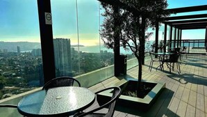 Eine Dachterrasse mit Glastisch und Stühlen, einem kleinen Baumkübel und Blick auf eine Stadtlandschaft.