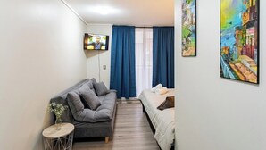 Ein kompakter Raum mit grauem Sofa, einem kleinen runden Beistelltisch, einem Bett mit weißer Bettdecke und einem an der Wand montierten Fernseher.