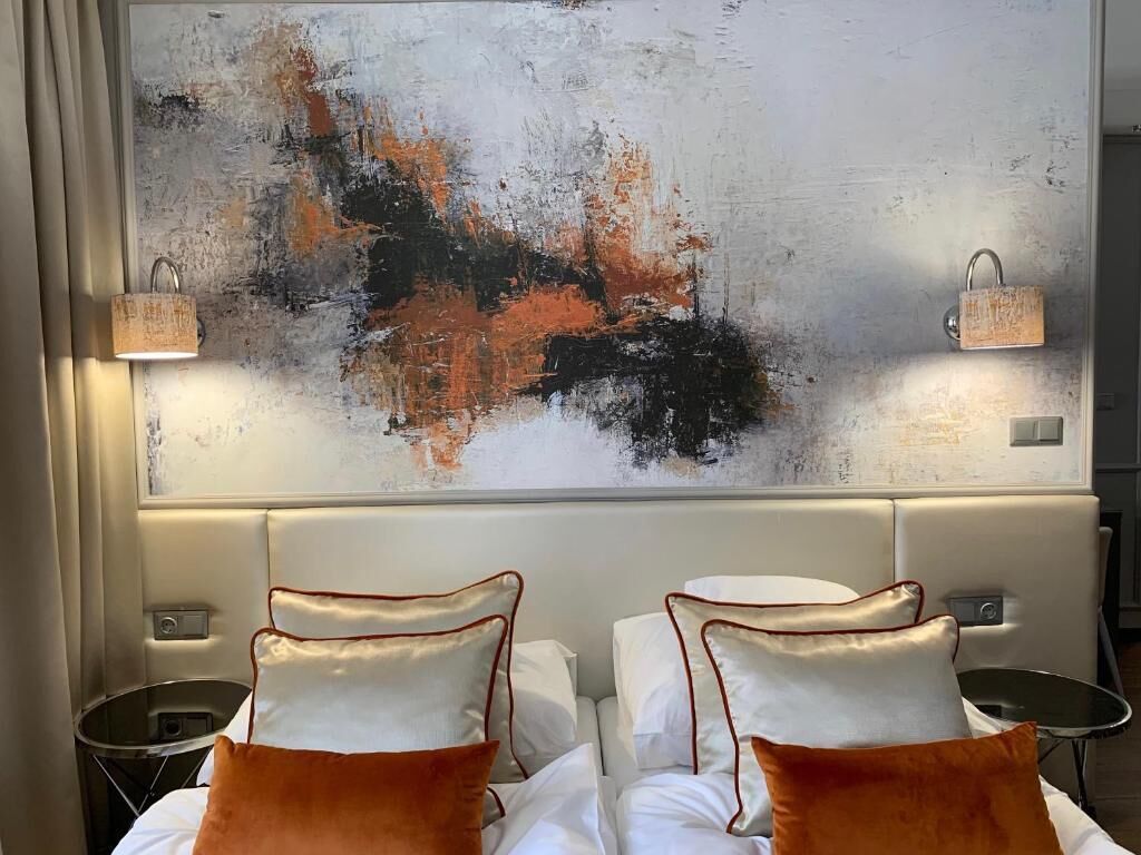A hotel room with a large abstract painting on the wall, two beds with white linens and orange pillows, and bedside tables with lamps.