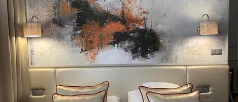 Habitación de hotel con un cuadro abstracto grande en la pared, dos camas con ropa blanca y almohadas naranjas, y mesitas de noche con lámparas.