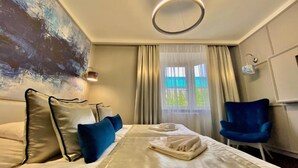 A hotel room with a bed, a blue chair, a painting on the wall, and a window with curtains. - Boutique Hotel Center (Kecskemét)
