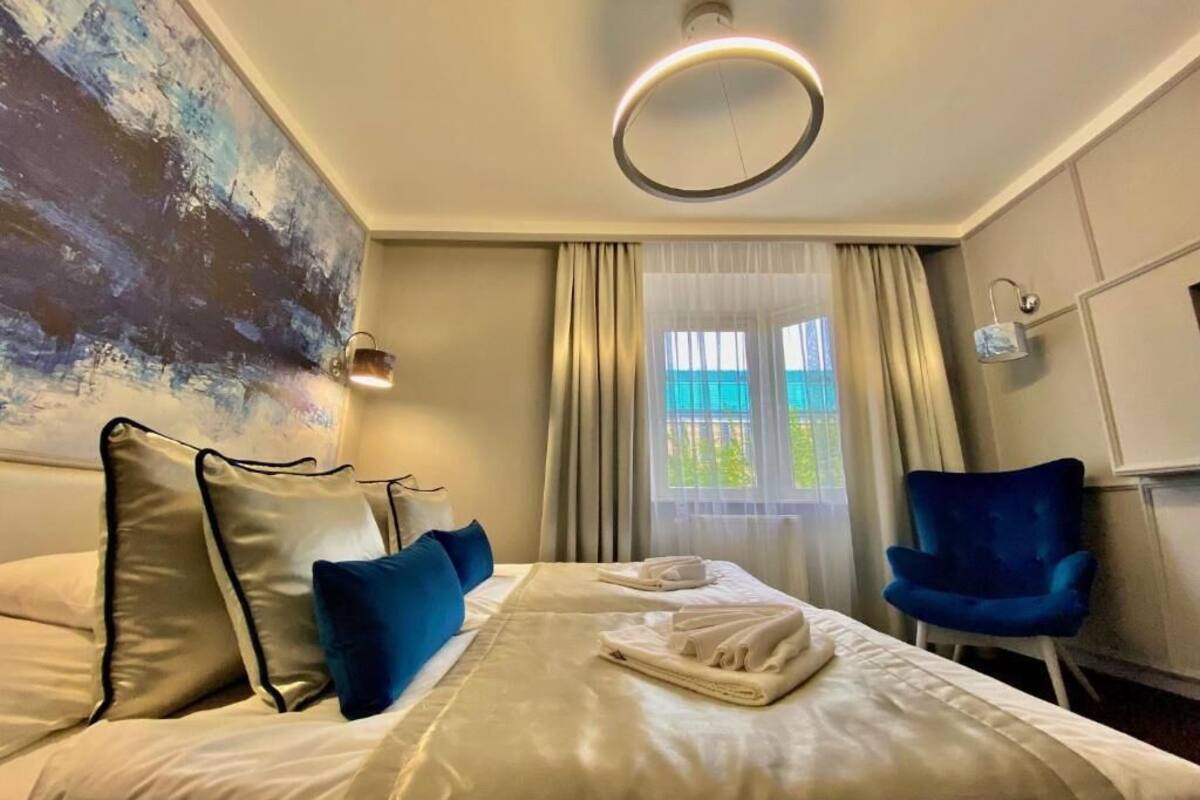 A hotel room with a bed, a blue chair, a painting on the wall, and a window with curtains.