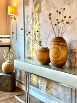 Un interior moderno con una mesa consola de cristal que sostiene tres jarrones de madera con plantas secas.