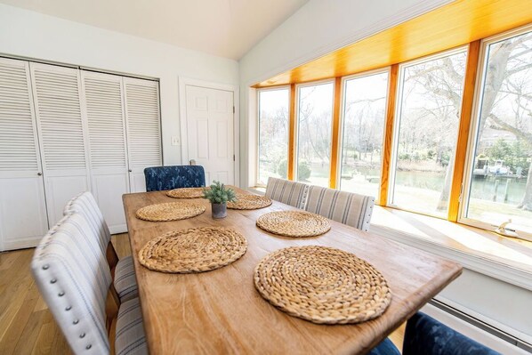 Comedor con mesa de madera, individuales tejidos, sillas acolchadas y vistas amplias a través de grandes ventanales.