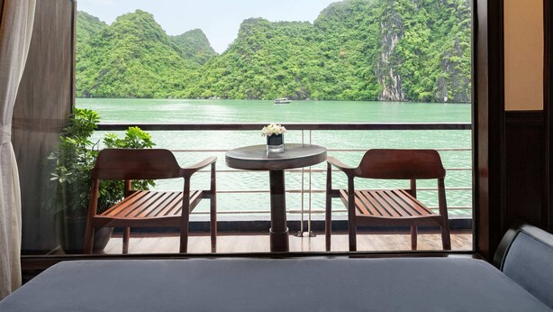 A balcony with two wooden chairs and a small table, overlooking a body of water with a boat and lush green hills in the background.