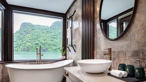 A bathroom with a bathtub, a sink, and a large mirror. There is a view of a forested area through the window.
