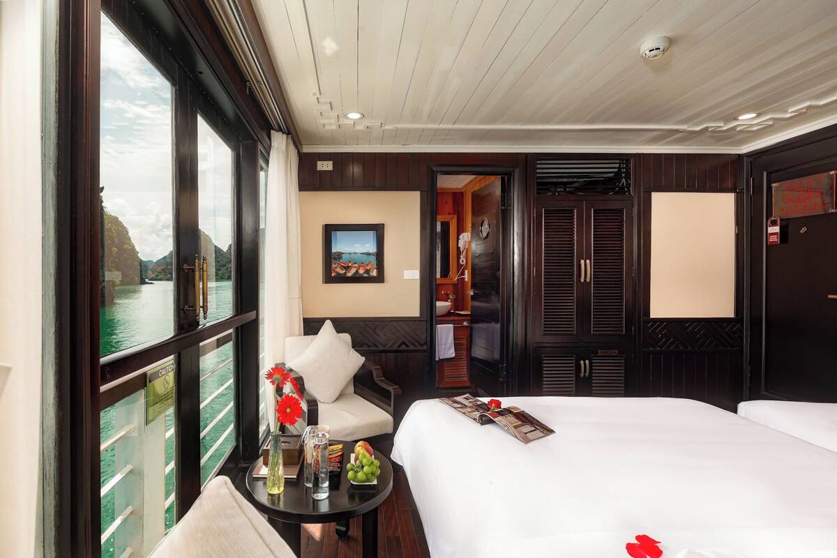 A hotel room with a large bed, a small table with a vase and fruit, a chair, a painting on the wall, and a view of a body of water through large windows.