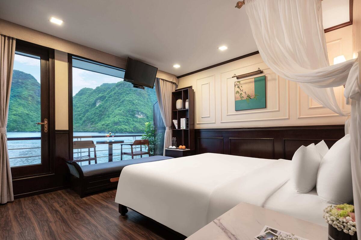 A hotel room with a large bed, a TV, a balcony with a view of a lake and mountains, and a dining area with a table and chairs.