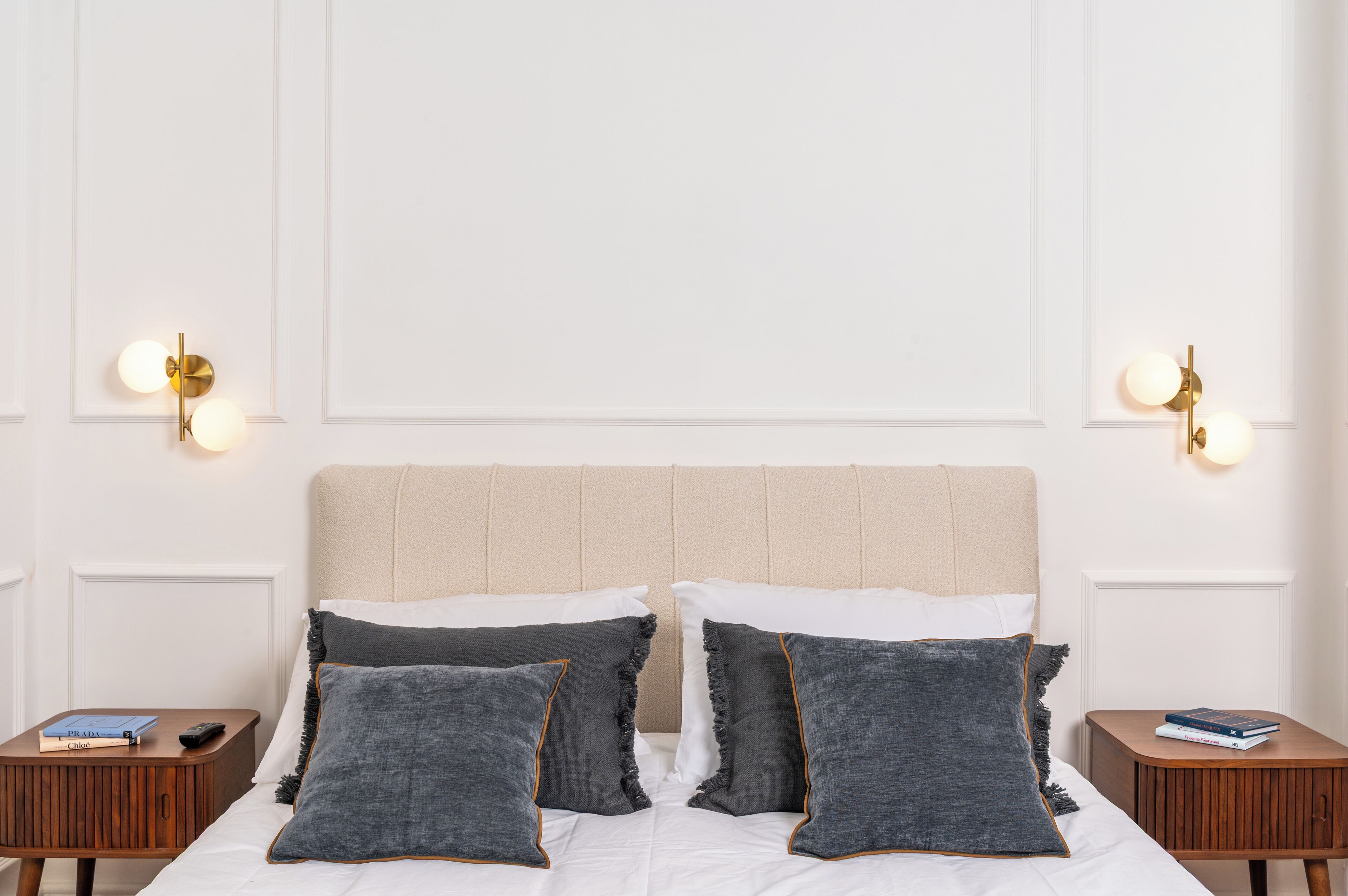 A neatly made bed with a beige headboard, two bedside tables with books and a remote control, and wall-mounted light fixtures.