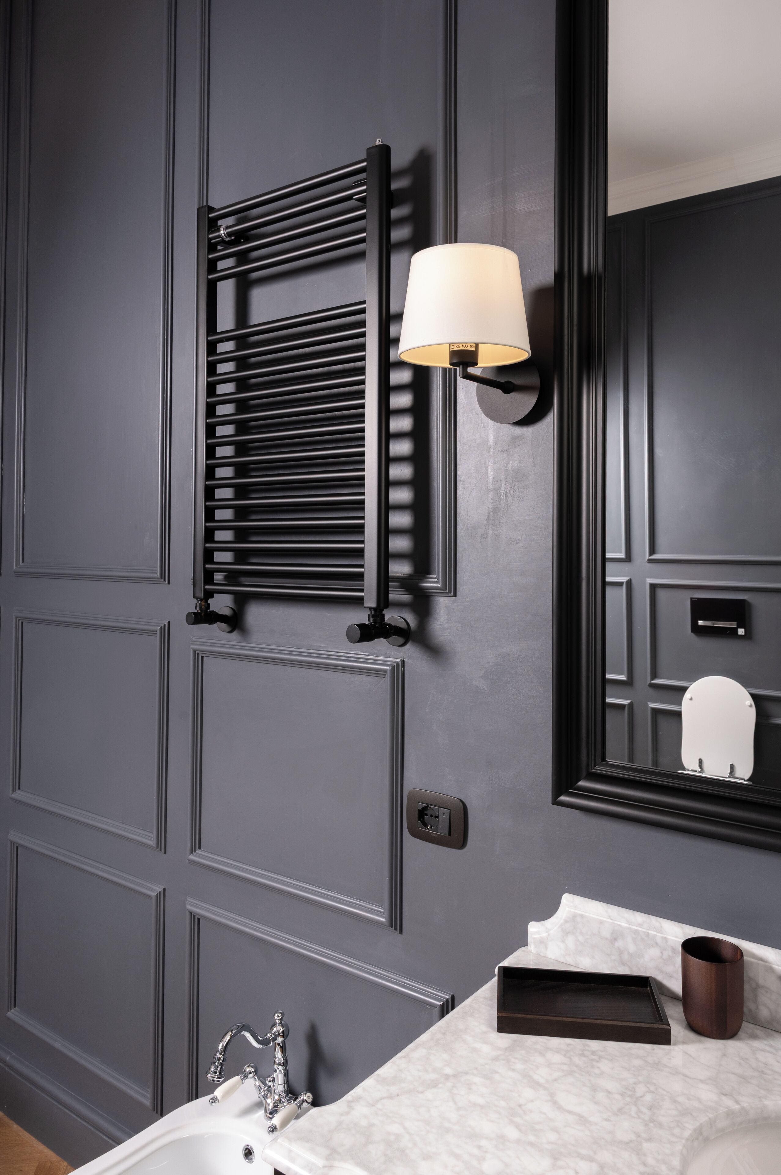 A bathroom with a dark grey wall, a towel rack, a wall-mounted lamp, a mirror, a sink, and a marble countertop.