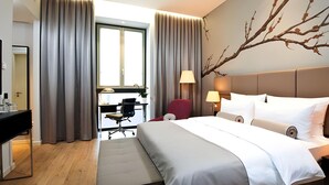 A hotel room with a bed, bedside tables, a desk, a chair, and a large window with curtains. - Hotel Berlin Potsdamer Platz by Leonardo Hotels (Berlin)