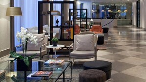 A modern hotel lobby with a checkered floor, seating area with chairs and ottomans, a glass coffee table with books, a bookshelf, and a decorative plant. - Hotel Berlin Potsdamer Platz by Leonardo Hotels (Berlin)