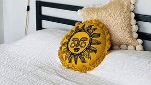 Un lit recouvert d’une couverture blanche à texture, doté d’une tête de lit noire et agrémenté de coussins décoratifs, dont un coussin jaune en forme de soleil et un coussin beige orné de pompons.