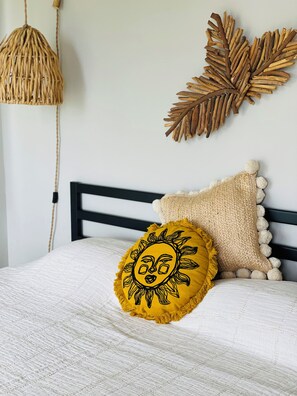 Een bed met een wit geweven beddekleed, een zwart hoofdbord en decoratieve kussens, waaronder een geel kussen in de vorm van een zon en een beige exemplaar met pompons.