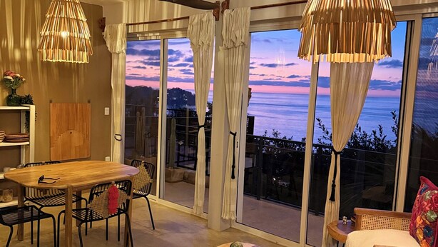 Un vaste espace de vie comprenant une table à manger en bois, des chaises noires, un canapé et une vue sur l’océan au coucher du soleil.