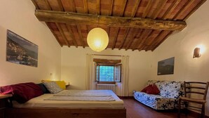 A bedroom with a wooden bed, a sofa, a chair, a window with curtains, and a hanging light fixture.