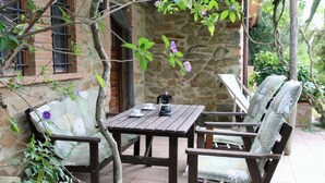 An outdoor patio with a wooden table and two chairs, a coffee maker, and a stone wall in the background.