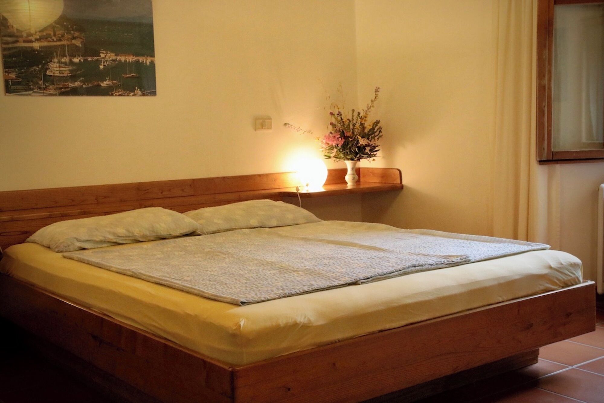 Ein Bett mit Holz Kopfteil, ein Nachttisch mit Lampe und ein an der Wand befestigtes Regal mit Blumen.