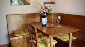 A wooden dining table with two wine glasses and a bottle of wine, a bench with cushions, and a wall-mounted shelf with dried flowers.
