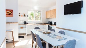 A modern kitchen with white cabinets, a dining table with chairs, a TV mounted on the wall, and a window with a view of trees. - Villa Delphi (Swieqi)