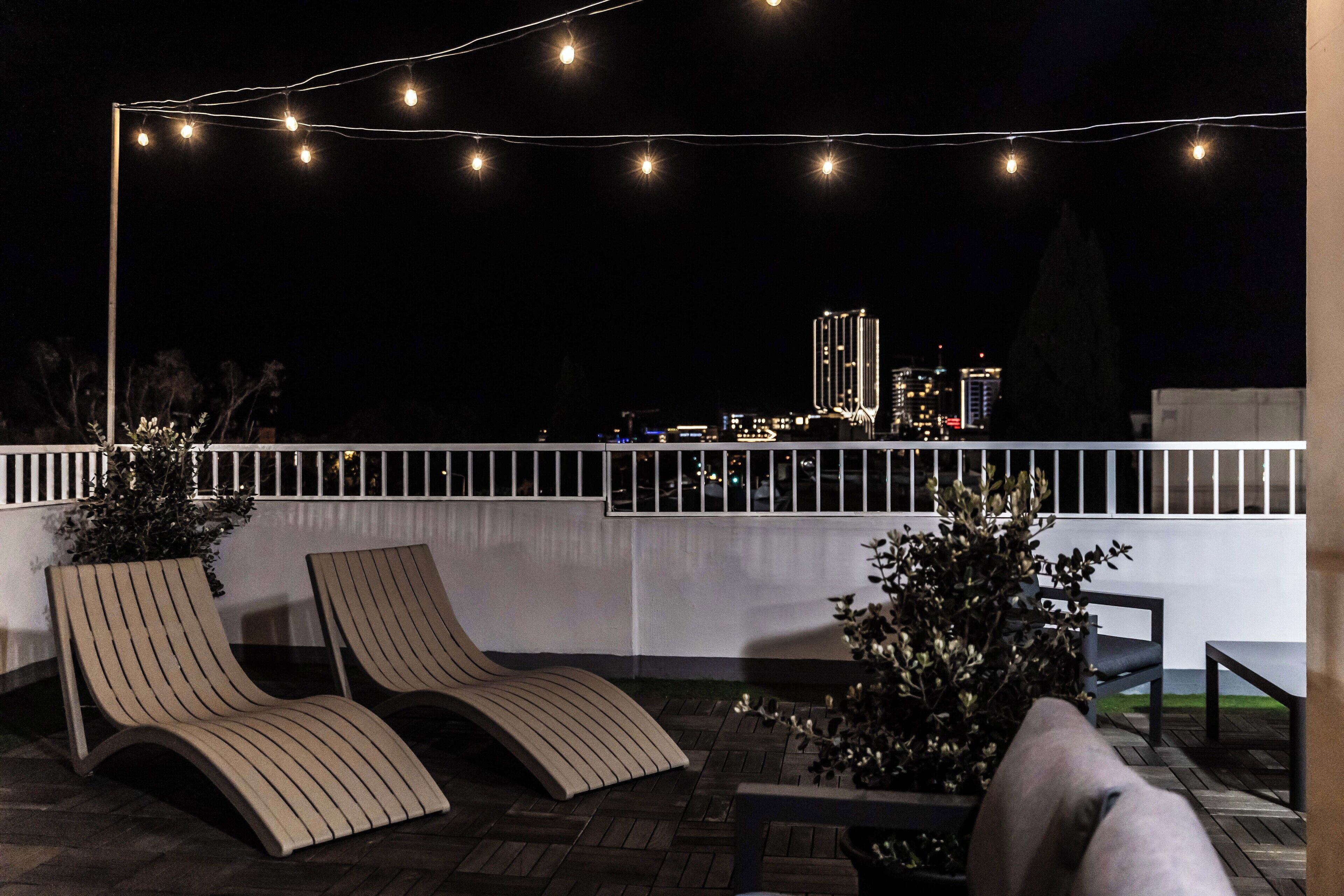 Una terraza en la azotea con dos sillones, luces colgantes y vistas a un paisaje urbano nocturno.