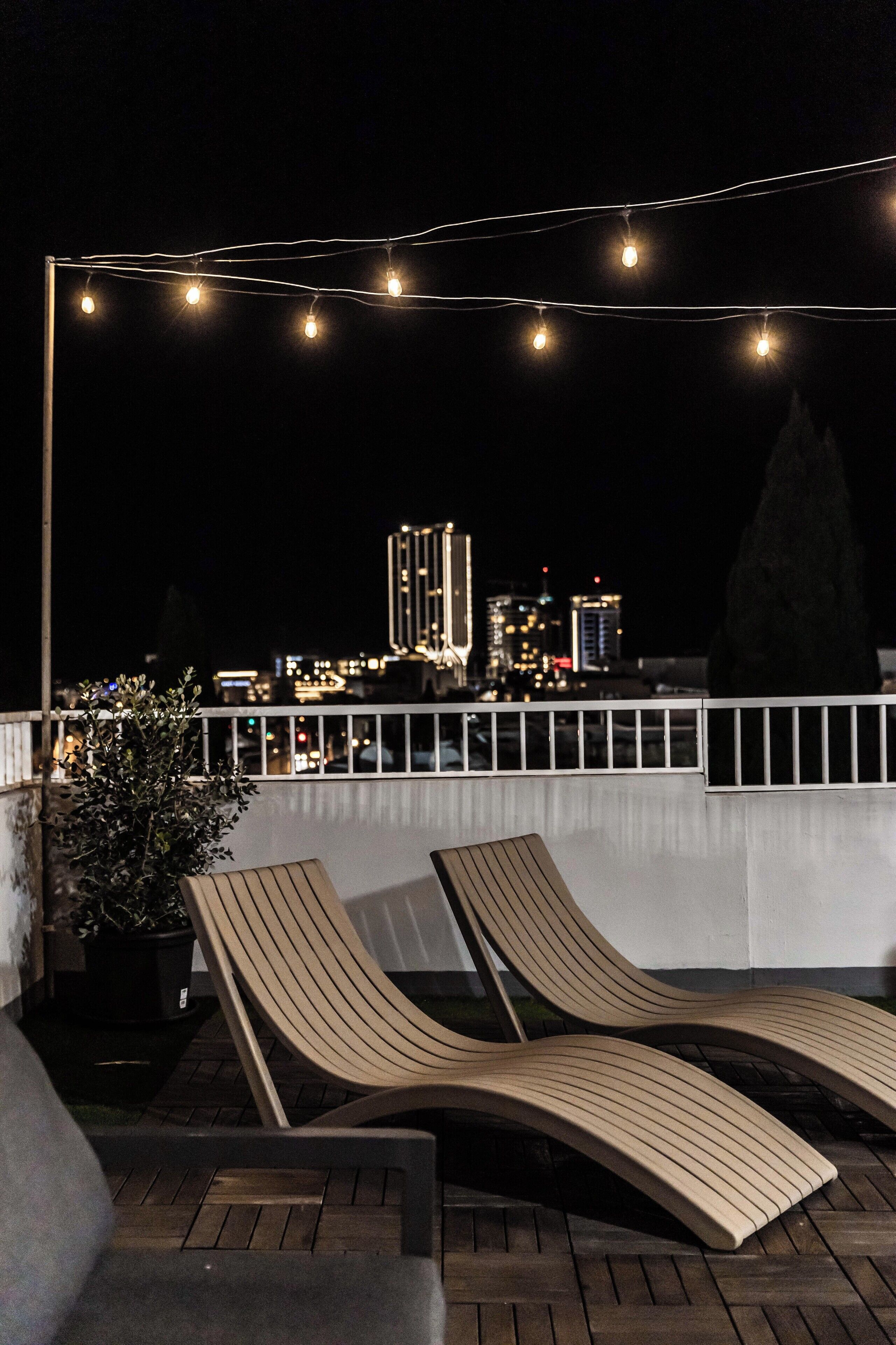 Une terrasse sur le toit, avec des chaises longues, des guirlandes lumineuses et une vue sur le paysage urbain en arrière-plan.