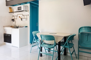 Uma cozinha compacta com micro-ondas, pia e lava-louças, uma mesa de jantar azul-turquesa com cadeiras, e uma porta azul.