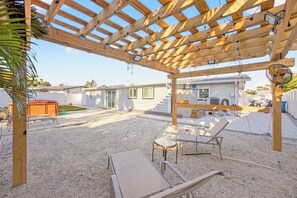 Een overdekt terras buiten, met een houten pergola, een zwembad en een huis op de achtergrond.