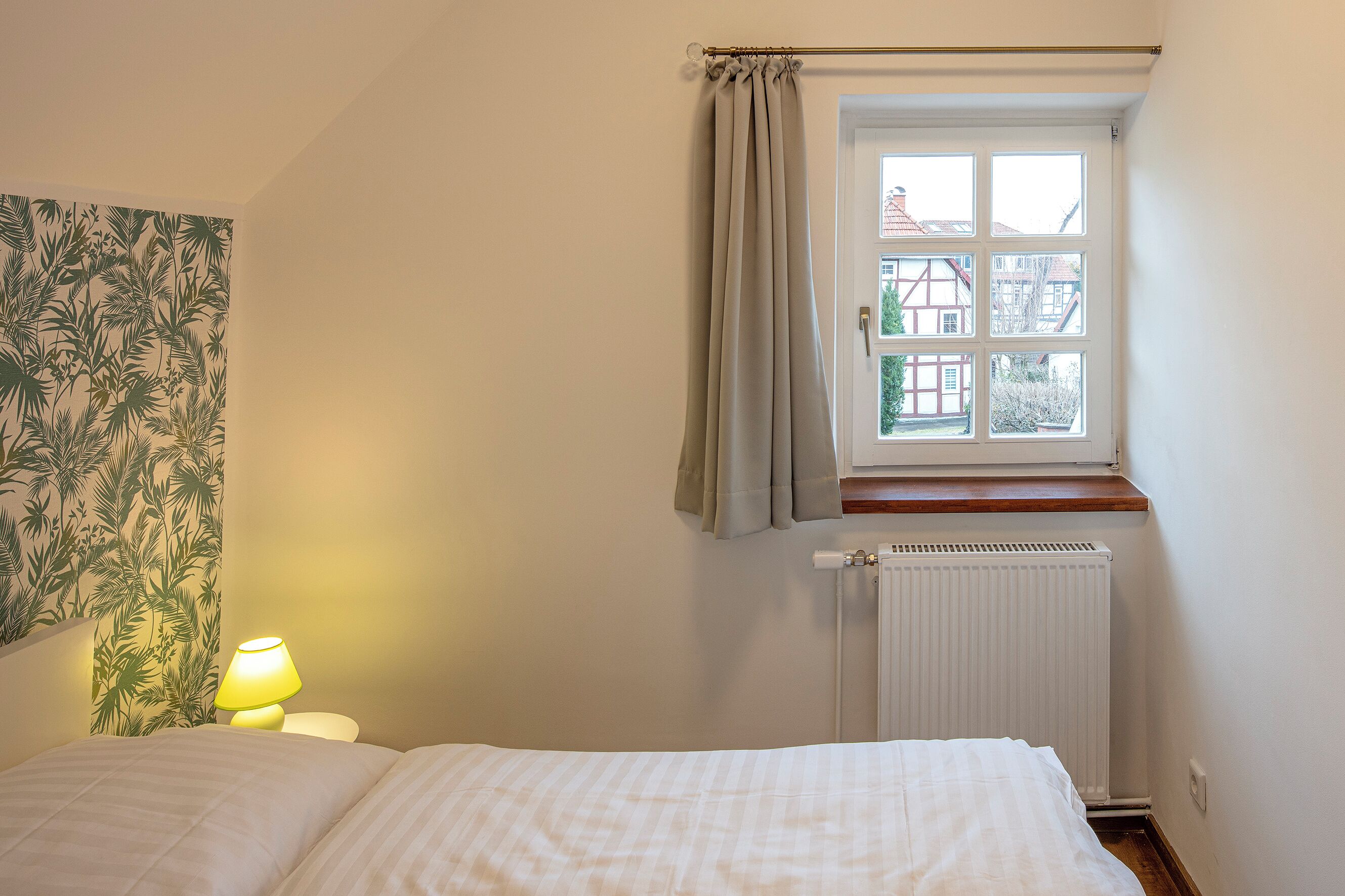 Une chambre à coucher avec un lit, une table de chevet avec une lampe, une fenêtre avec des rideaux et un radiateur.