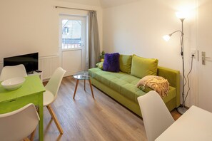 Ein modernes Wohnzimmer mit einem grünen Sofa, weißen Stühlen, einem Glastisch und einem Fernseher.