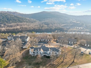 A residential area with multiple houses, a large central house with a prominent porch, and a lake in the background. - Pointe Royale | 2-Car Garage | Pools | Golf (Branson)