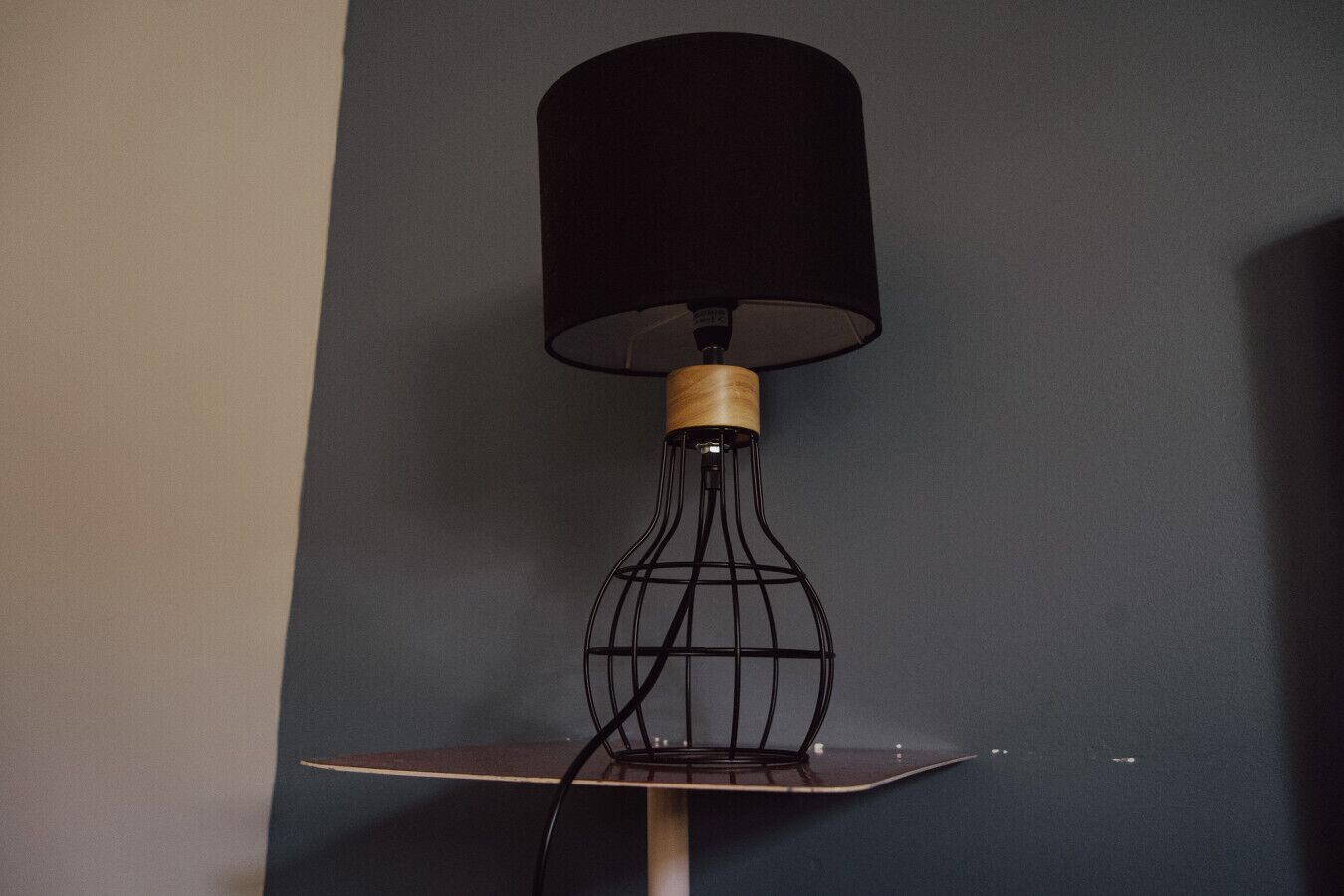 Une lampe de sol moderne, dotée d’un abat-jour noir et d’un pied en bois, placée sur une petite table adossée à un mur sombre.
