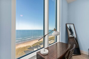 Ein Zimmer mit großem Fenster mit Blick auf einen Strand, einem Schreibtisch mit Lampe und einem an der Wand lehnten Spiegel.