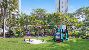 Ein Spielplatz mit Schaukeln und einer Kletterkonstruktion, umgeben von Palmen und einem Gebäude im Hintergrund.