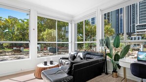 Ein modernes Wohnzimmer mit einem schwarzen Ledersofa, einem runden Holztisch und einem Laptop auf einem Schreibtisch. Große Fenster bieten Blick auf einen Garten und eine Stadtansicht.