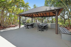 An outdoor patio with tables and chairs, surrounded by lush greenery and a covered area with a tiled roof. - Loft w/ Glass Ceiling, Pool & Tennis, Surfers (Surfers Paradise)